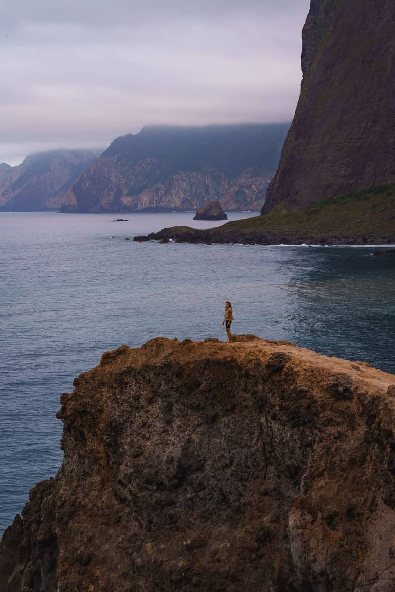 A lone figure standing on a volcanic sea cliff above the Atlantic, misty mountains in the distance, Madeira.